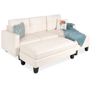 This is an L-shaped sectional sofa with an ottoman in white upholstery, featuring tufted cushioning, black tapered legs, and adorned with two patterned pillows and a throw blanket.