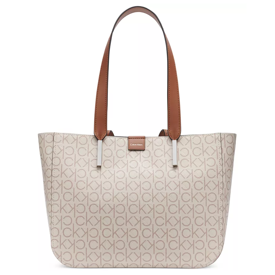 Calvin Klein Logo Medium Tote features beige fabric with an all-over \“CK\“ pattern, brown leather handles, and a small Calvin Klein logo tag at the top center.