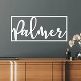 A modern wall sign featuring the word \“Palmer\“ in a script font within a rectangular frame is displayed on a gray wall above a wooden cabinet.