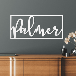A modern wall sign featuring the word \“Palmer\“ in a script font within a rectangular frame is displayed on a gray wall above a wooden cabinet.