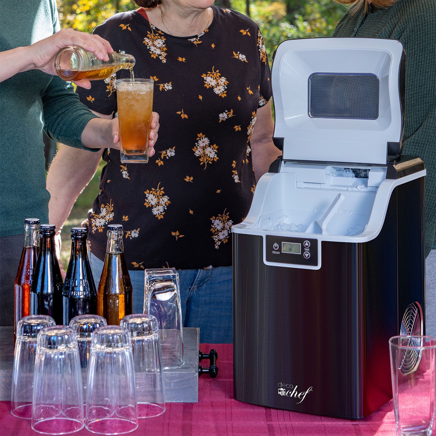 A portable ice maker is displayed alongside various sized glass bottles and empty glasses, with a person pouring a beverage into a glass.