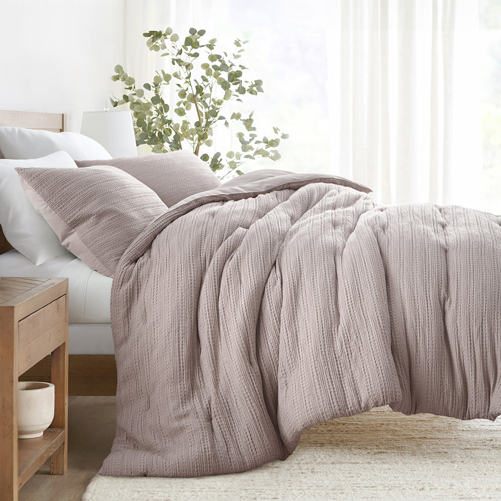 A cozy bed setup featuring a textured, light mauve comforter with two matching pillows, complemented by a wooden nightstand and a ceramic planter.