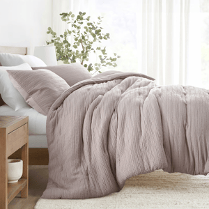 A cozy bed setup featuring a textured, light mauve comforter with two matching pillows, complemented by a wooden nightstand and a ceramic planter.