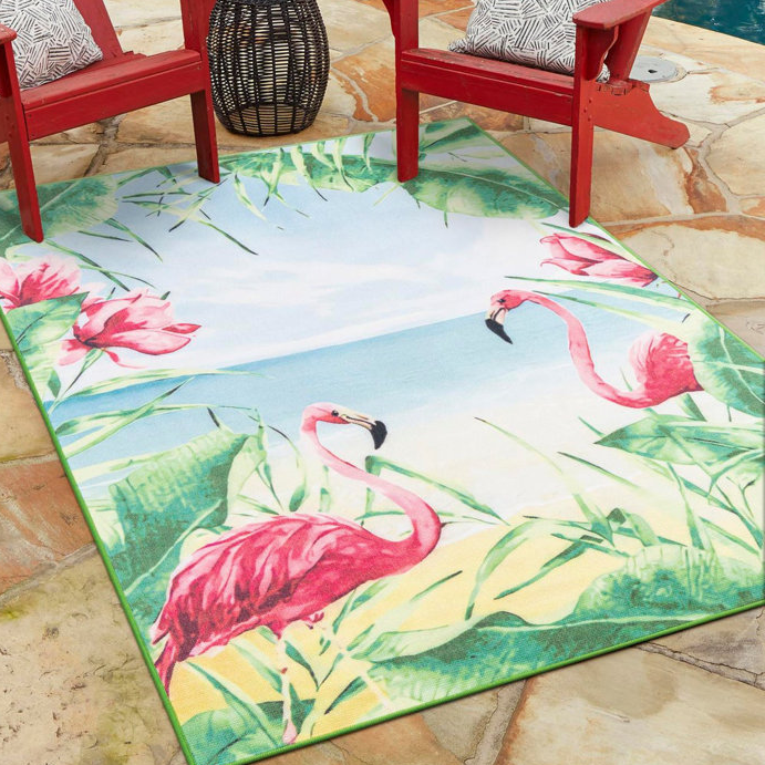 9' x 12' outdoor rug featuring a tropical scene with pink flamingos and green foliage on a beach background, placed under red Adirondack chairs on a stone patio.