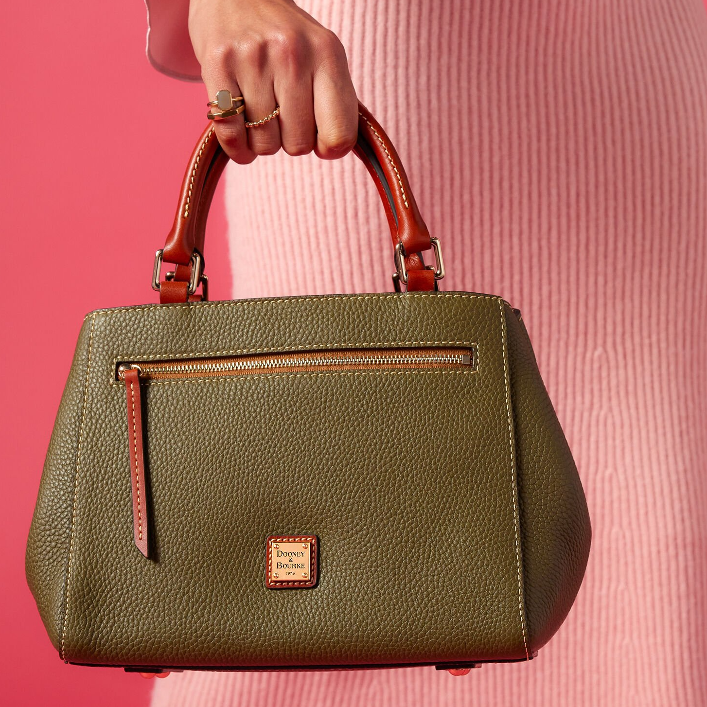 A woman holds a Dooney & Bourke olive green pebbled leather handbag with tan handles and a front zipper pocket, set against a pink background.