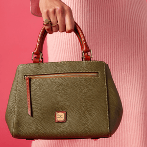 A woman holds a Dooney & Bourke olive green pebbled leather handbag with tan handles and a front zipper pocket, set against a pink background.