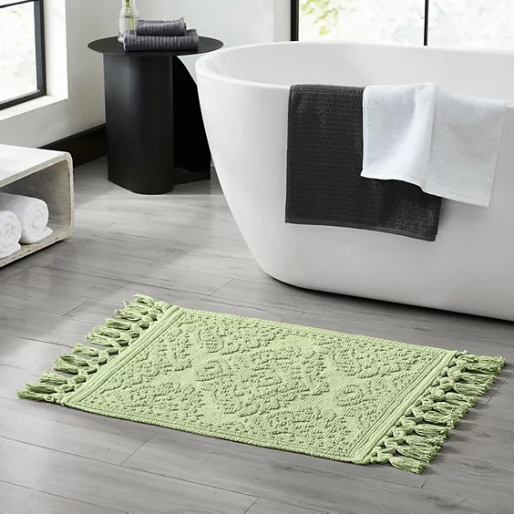 A light green bath mat with tassel edges on both sides is placed on a gray tiled bathroom floor near a white bathtub. A dark gray towel and a white towel hang on the bathtub, with a small side table and rolled towels nearby.