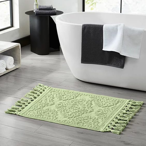 A light green bath mat with tassel edges on both sides is placed on a gray tiled bathroom floor near a white bathtub. A dark gray towel and a white towel hang on the bathtub, with a small side table and rolled towels nearby.