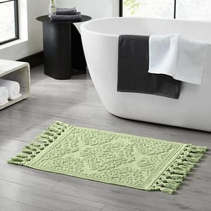A light green bath mat with tassel edges on both sides is placed on a gray tiled bathroom floor near a white bathtub. A dark gray towel and a white towel hang on the bathtub, with a small side table and rolled towels nearby.