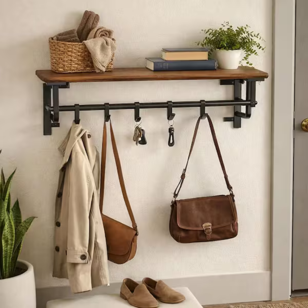 A wall-mounted shelf and hook rack features a wooden shelf holding books and a plant, and hooks below holding a coat, handbags, and keys. Beneath it sits a pair of shoes.
