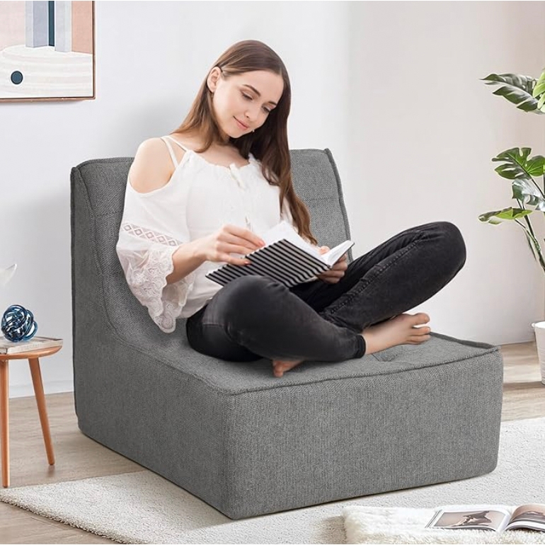 A woman sits on a gray 39-inch foam chair in a modern living room. She is reading a notebook, dressed in a white off-shoulder blouse and black pants, surrounded by a small table, a plant, and decor elements.