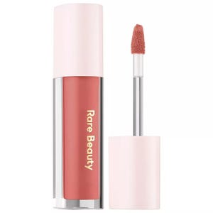 Rare Beauty Liquid Eyeshadow features a sleek tube with a soft pink cap, containing a coral shade. The product includes a doe-foot applicator for easy application.