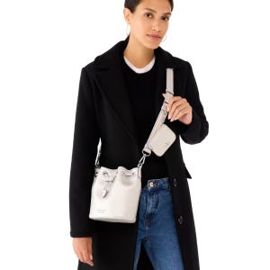 The Kate Spade Bucket Bag Set features a small white bucket bag with a drawstring closure and an adjustable strap, accompanied by a matching cardholder. The bag is styled with a chic black coat and jeans.