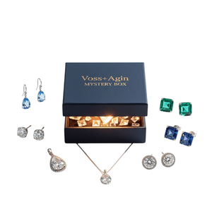 A Voss+Agin Mystery Box is surrounded by elegant jewelry, including blue teardrop earrings, clear and blue stud earrings, emerald-green square earrings, a teardrop pendant, and a round pendant necklace.