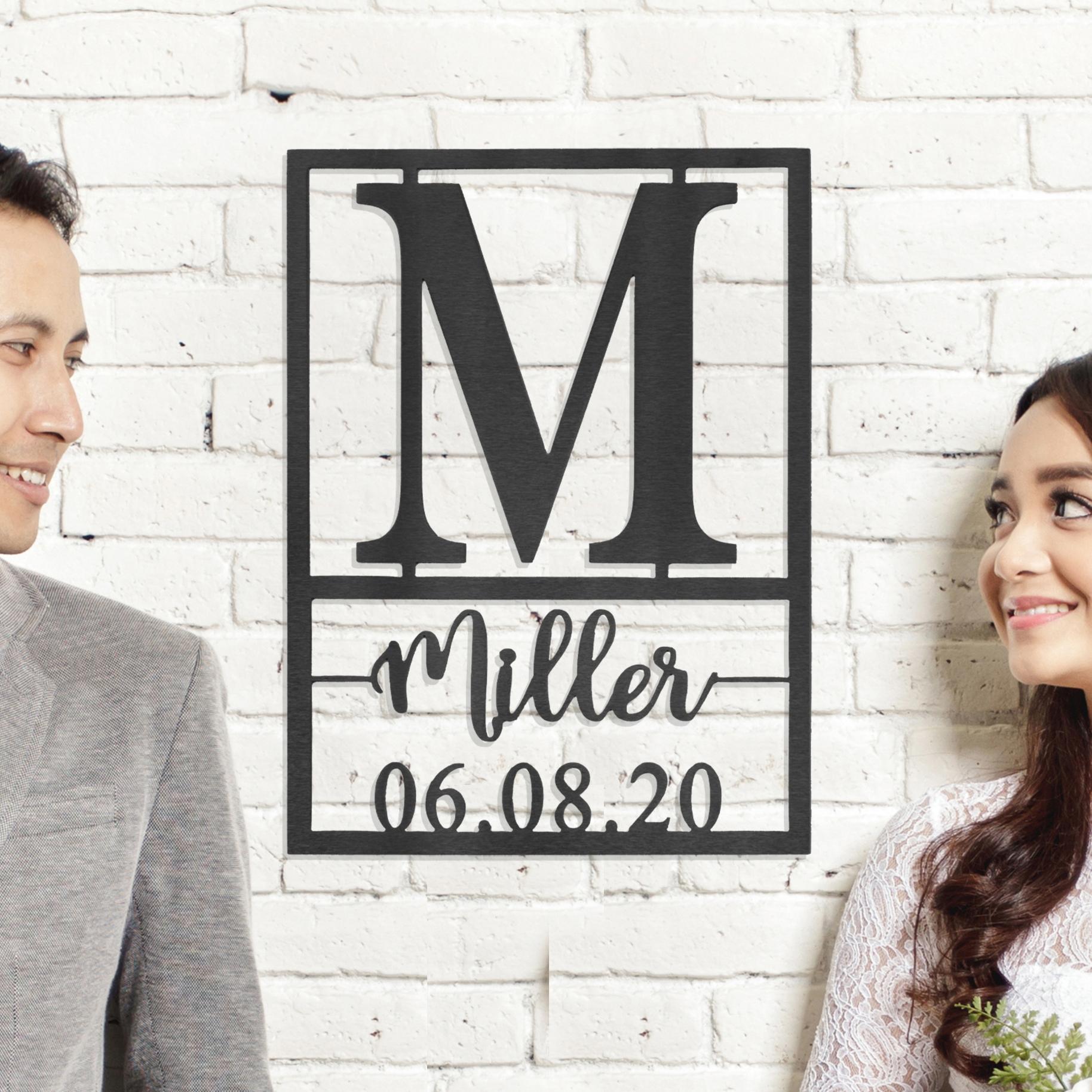 A personalized wall art featuring a large initial 'M' with the name 'Miller' and the date '06.08.20'. Two people are smiling beside it.