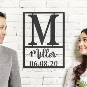 A personalized wall art featuring a large initial 'M' with the name 'Miller' and the date '06.08.20'. Two people are smiling beside it.