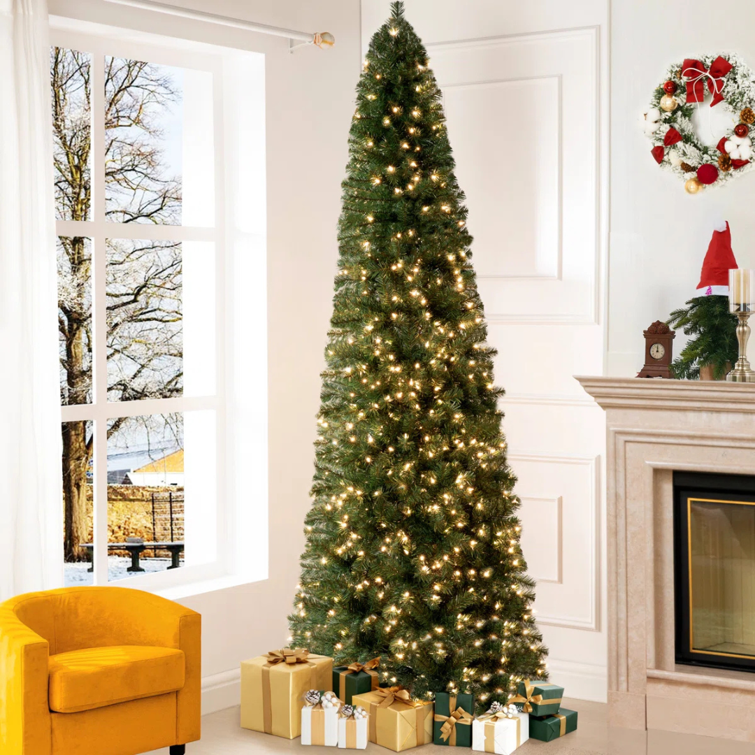 A tall, slim Christmas tree with warm white lights is surrounded by wrapped gifts. The scene includes an indoor setting with a fireplace, a wreath, and a yellow chair against a window view of a snowy outdoor landscape.