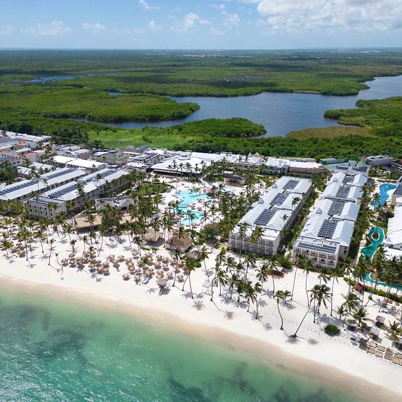 A beachfront resort with multiple pools, palm trees, and thatched umbrellas, situated next to clear turquoise waters and lush green surroundings.