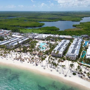 A beachfront resort with multiple pools, palm trees, and thatched umbrellas, situated next to clear turquoise waters and lush green surroundings.