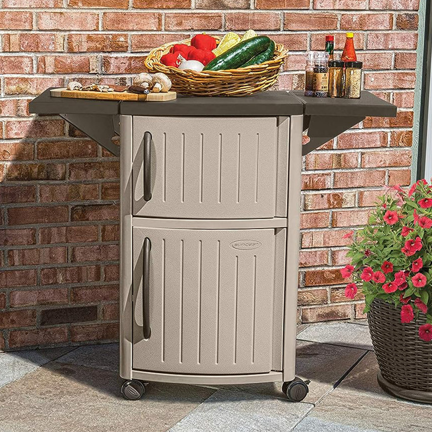 A Suncast serving cart with two cabinets and an extendable top shelf, mounted on wheels, positioned on a patio alongside a brick wall.