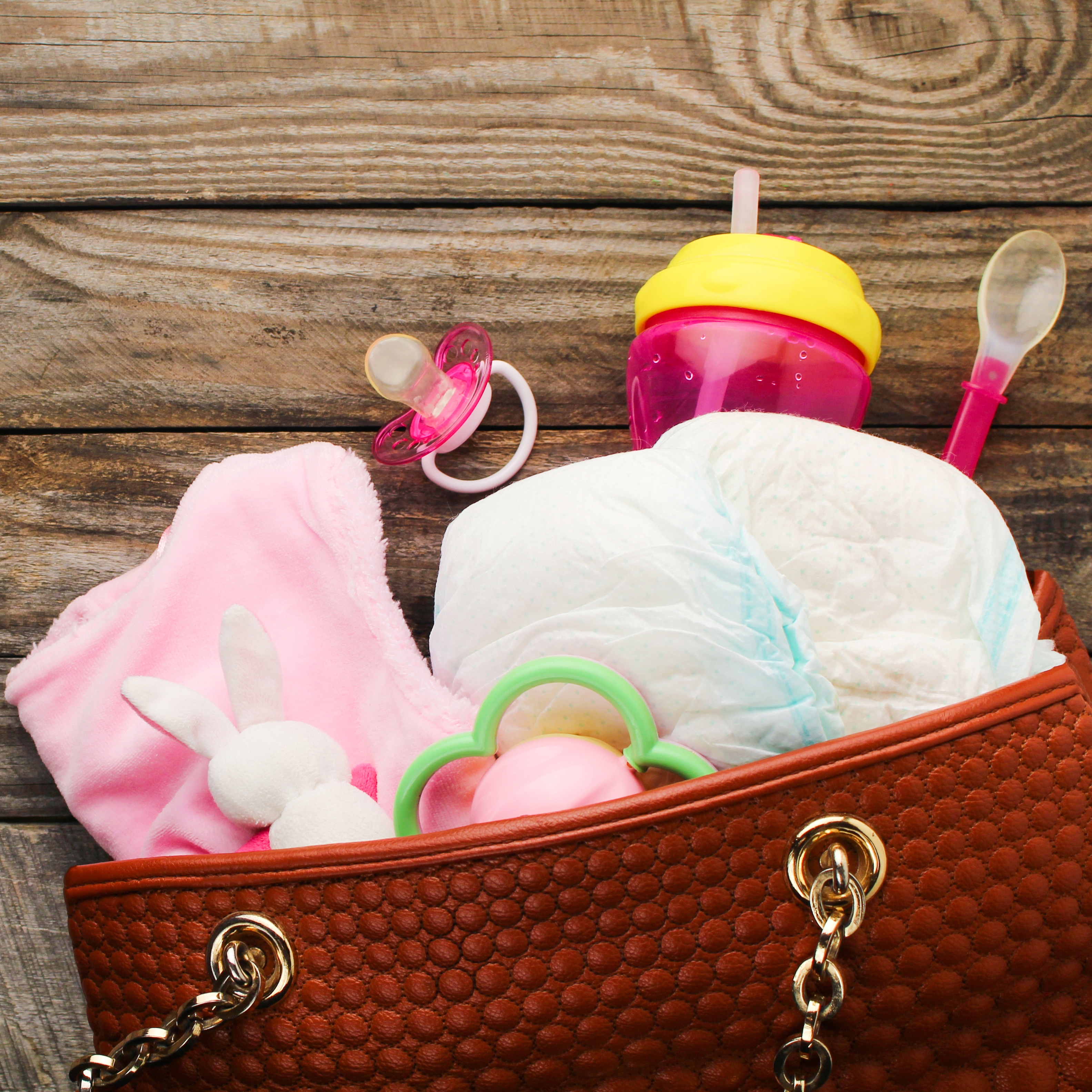 A brown diaper bag with baby essentials: diapers, pacifier, sippy cup, spoon, plush bunny, and pink blanket.