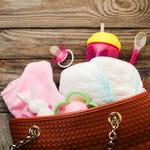 A brown diaper bag with baby essentials: diapers, pacifier, sippy cup, spoon, plush bunny, and pink blanket.