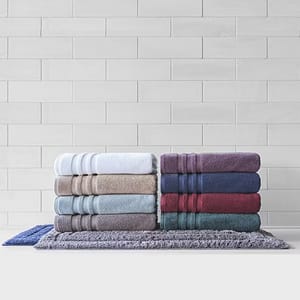 Stacked towels in various colors, including white, beige, light blue, purple, navy, and burgundy, displayed on textured mats against a tiled wall. Made from Egyptian cotton, they appear plush and neatly arranged.