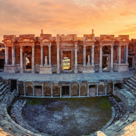 An ancient Roman amphitheater with stone seating and a detailed, columned stage, set against a dramatic sunset sky.