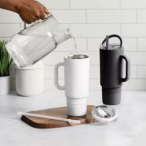 Two insulated travel mugs, one white and one black, are shown; one is being filled with water, both have lids and straws, and they rest on a wooden board.