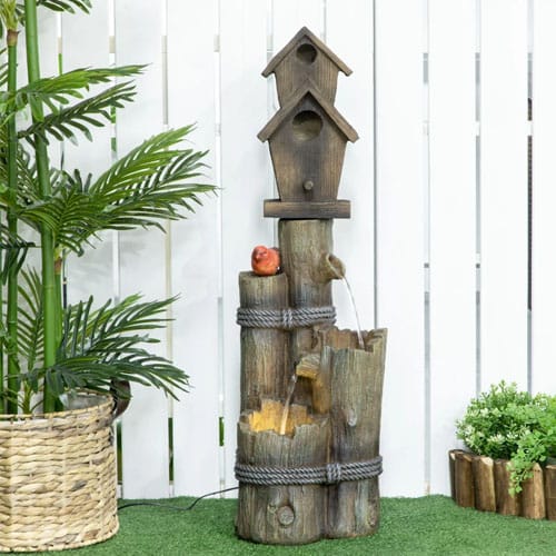 Wooden birdhouse fountain with a faux bird perched on the side, set against a white fence with greenery.