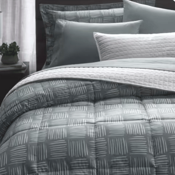 A bed is shown with a modern geometric-patterned comforter, accompanied by matching pillows and a quilted white throw, against a muted background featuring a nightstand with a lamp and a small plant.