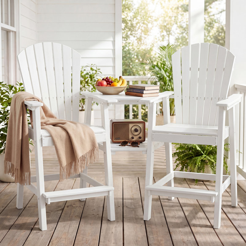 White Adirondack chairs with a connecting table are on a wooden porch, adorned with a beige throw, radio, books, sunglasses, and a fruit bowl.