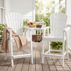 White Adirondack chairs with a connecting table are on a wooden porch, adorned with a beige throw, radio, books, sunglasses, and a fruit bowl.