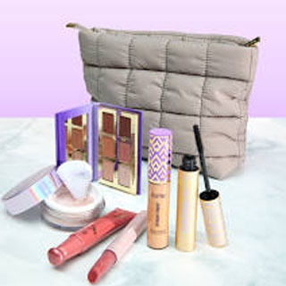 A quilted makeup bag with various cosmetics including a powder with puff, eyeshadow palette, concealer, mascara, and lip glosses, displayed on a marble surface against a purple background.