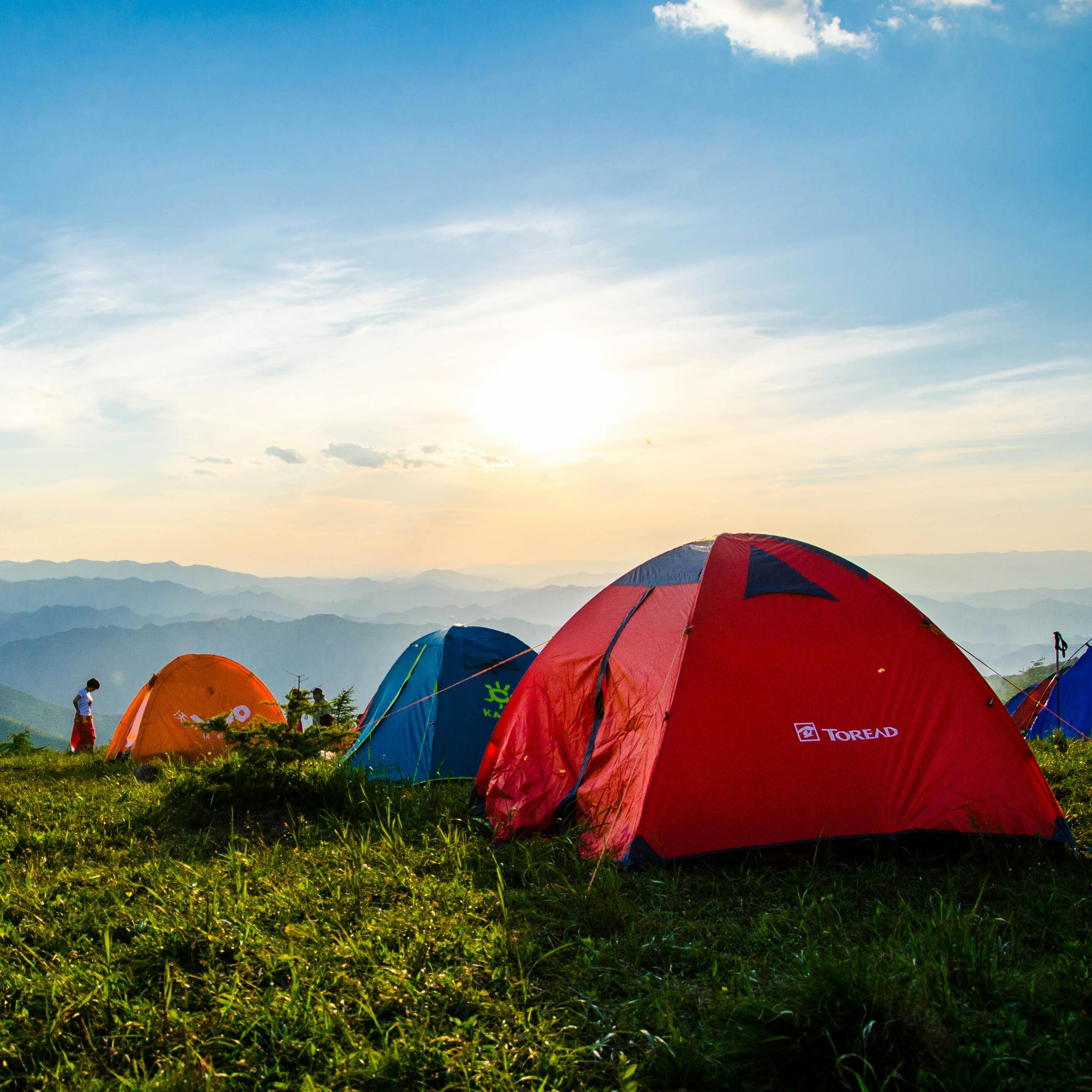 Several camping tents are set up on a grassy hill with a sunrise and mountain backdrop.