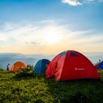 Several camping tents are set up on a grassy hill with a sunrise and mountain backdrop.