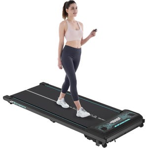 A woman is walking on a compact, foldable walking pad treadmill, featuring a slim design with a digital display and remote control. The treadmill is black with light blue accents and is suitable for indoor exercise.