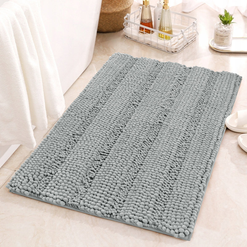 Light blue chenille bath mat with a textured, striped pattern, placed on a tiled bathroom floor near white towels, bath products, and white slippers.