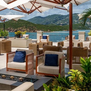 Outdoor seating area with wicker chairs, blue cushions, white umbrellas, and a view of the ocean and green hills in the background.