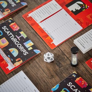 Classic Scattergories game includes pads with category lists, a 20-sided letter die, a sand timer, and game folders. Players roll the die to select a letter, then write words starting with that letter for each category before time runs out.