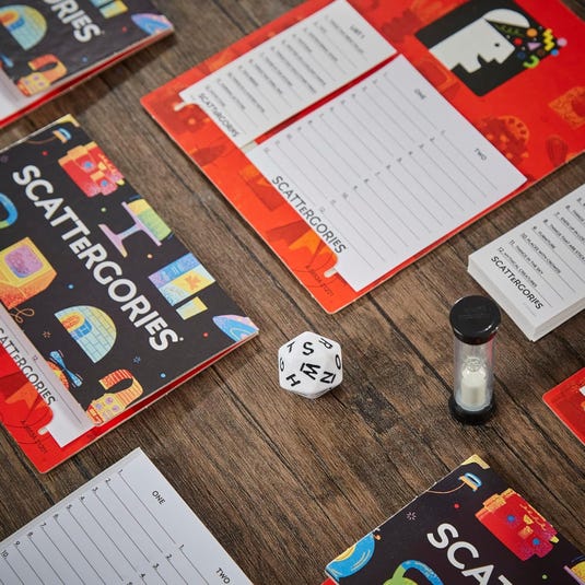Classic Scattergories game includes pads with category lists, a 20-sided letter die, a sand timer, and game folders. Players roll the die to select a letter, then write words starting with that letter for each category before time runs out.