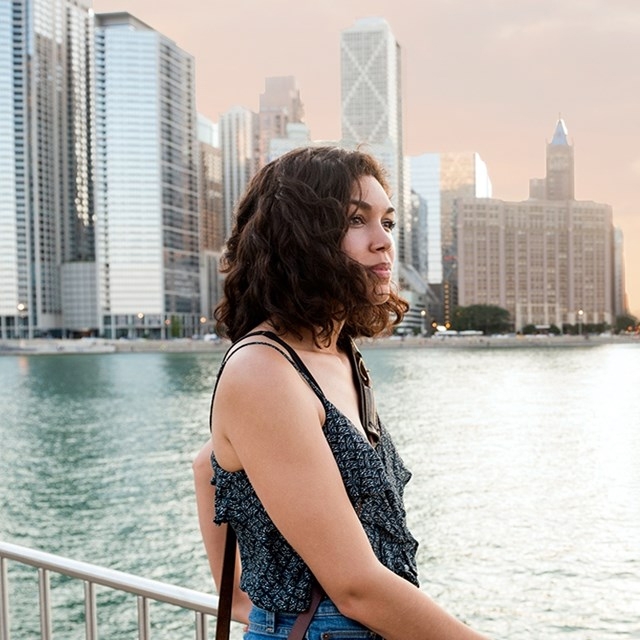 A woman is standing by a waterfront with a city skyline in the background. She is wearing a patterned sleeveless top and has a crossbody bag.