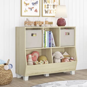 A wooden storage shelf with three cubbies, housing toys, books, and baskets, featuring a lamp and decorative items on top. Two framed pictures adorn the wall above, with a colorful rug and woven basket nearby.