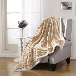 A beige, ribbed throw blanket with white trim is draped over a light gray armchair. Nearby, a small white table holds a vase with purple flowers, set against a window with sheer curtains.