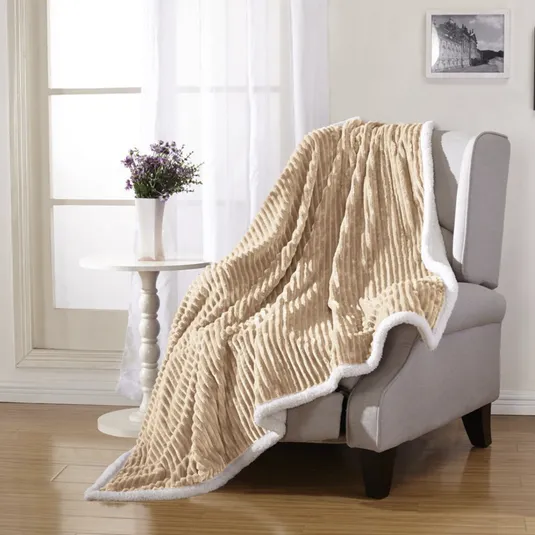 A beige, ribbed throw blanket with white trim is draped over a light gray armchair. Nearby, a small white table holds a vase with purple flowers, set against a window with sheer curtains.
