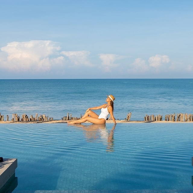 A person lounges on a submerged pool bench, wearing a white swimsuit and a straw hat, with the ocean and blue sky in the background.