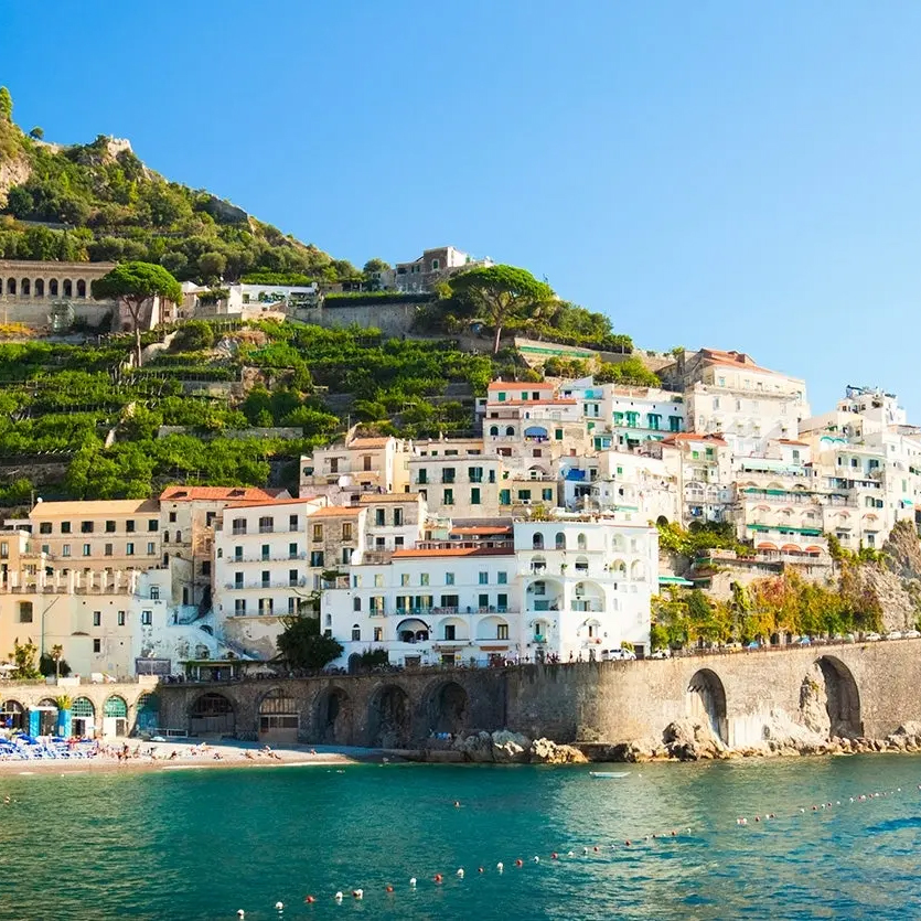 Coastal hillside town with white and pastel-colored buildings, lush greenery, and a stone arch promenade by the blue sea.
