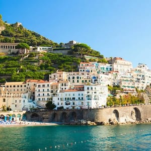 Coastal hillside town with white and pastel-colored buildings, lush greenery, and a stone arch promenade by the blue sea.