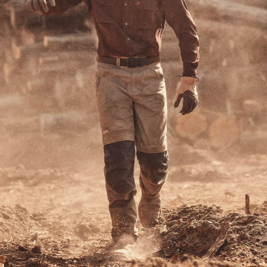 A man wearing a brown shirt and knee-padded pants, carrying a chainsaw while wearing a tan baseball cap and gloves, walks through a dusty, wood-filled area.