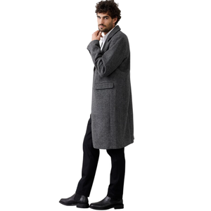 A man wearing a long, gray overcoat with black pants and black leather boots, standing against a white background.